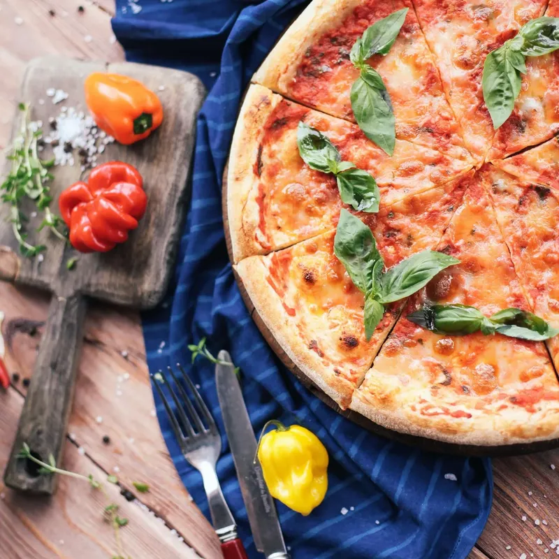 A sliced cheese pizza with basil on a wooden board beside a blue cloth and red peppers